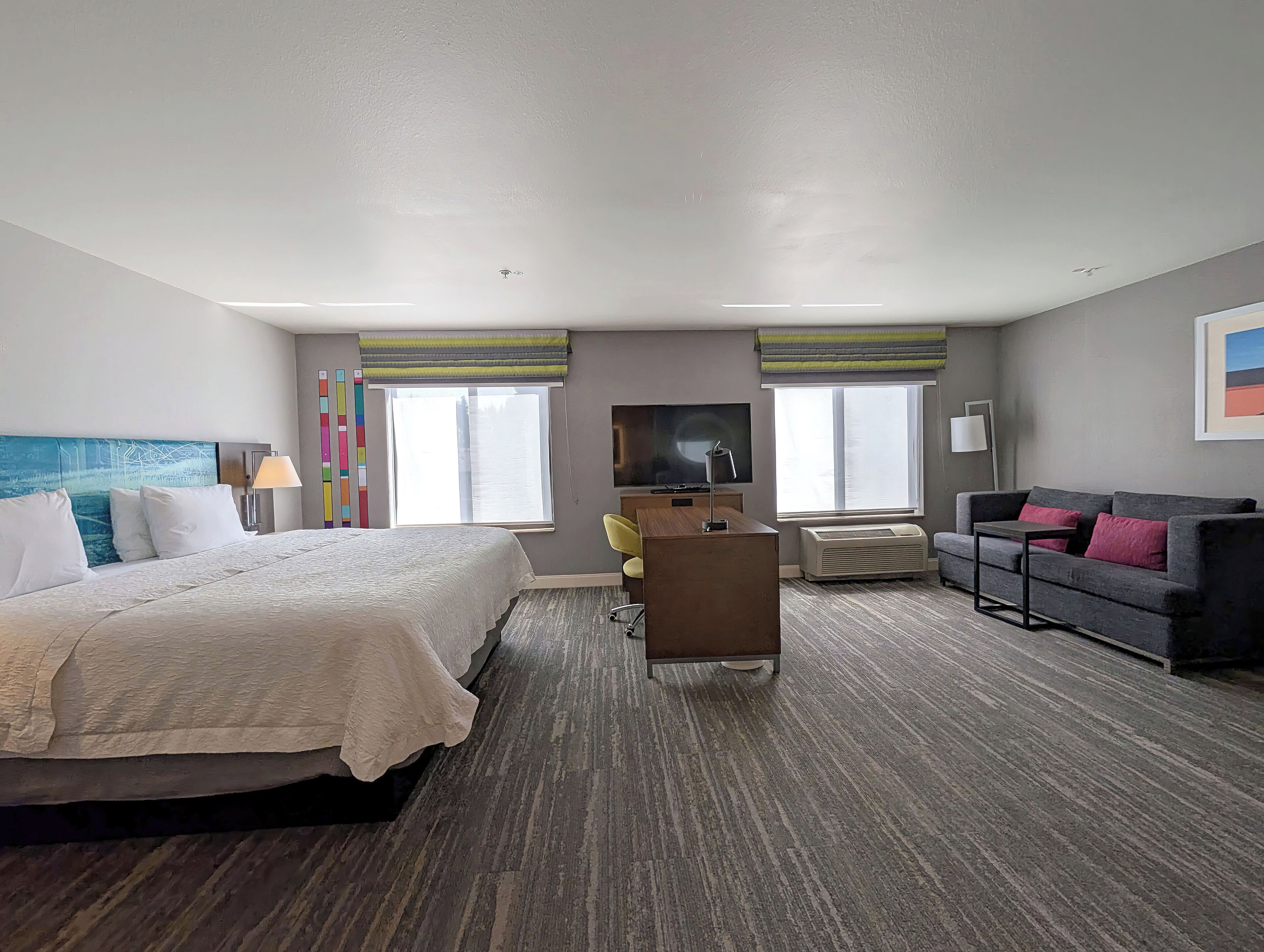 Una habitación de hotel con cama, escritorio, sofá y dos ventanas con cortinas.