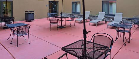 An outdoor patio area with metal tables and chairs, red umbrellas, and a beige wall.