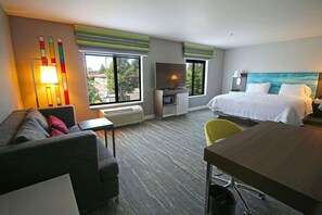 Hotellihuone, jossa on sänky, sohva, työpöytä, televisio ja ikkuna, josta on näkymä rakennuksiin ja puihin.