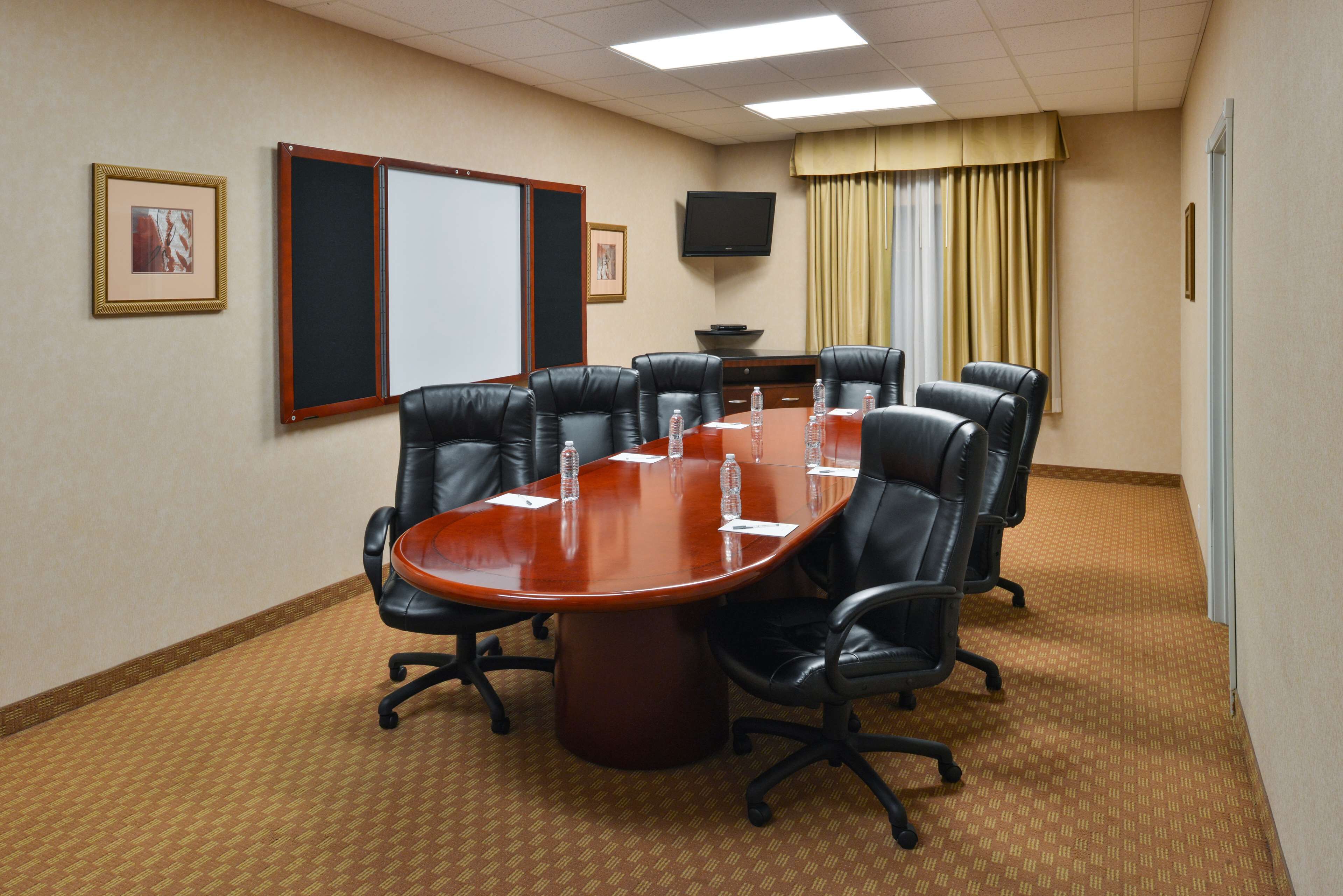 Un salón de conferencias con una gran mesa ovalada, seis sillas de cuero negro, una pizarra, un televisor de pantalla plana y un ventanal con cortinas.
