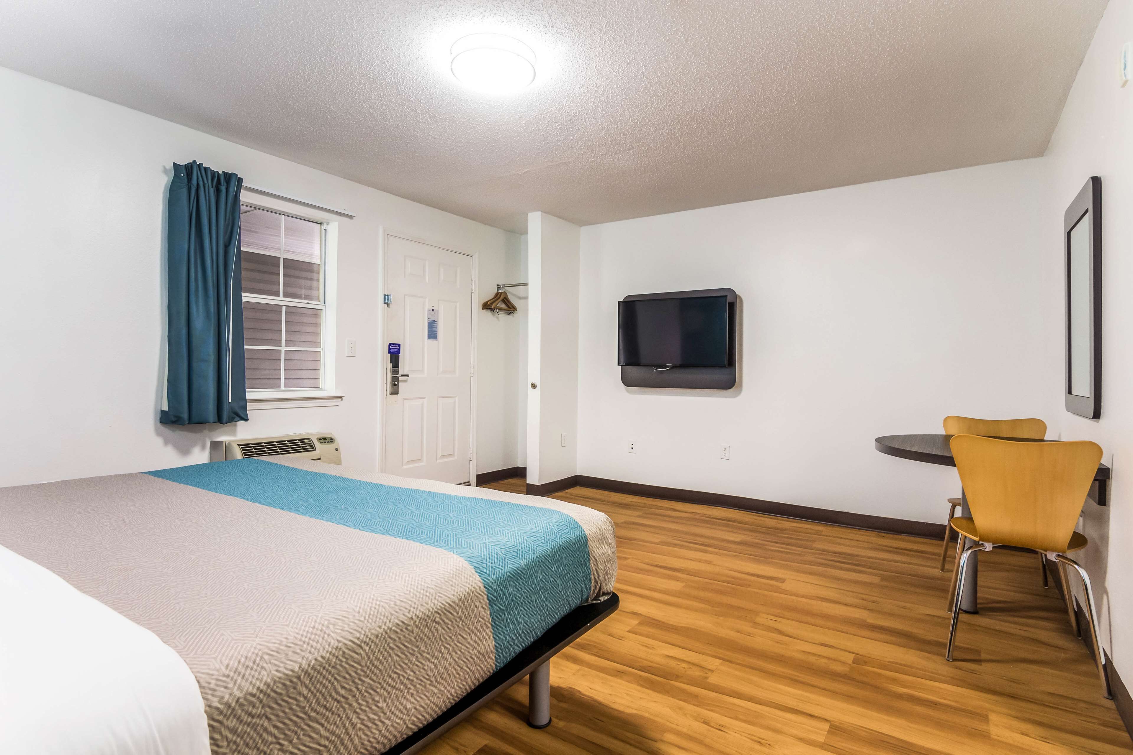 Habitación de hotel con cama, televisor montado en la pared, una mesita con una silla y una ventana con cortinas.