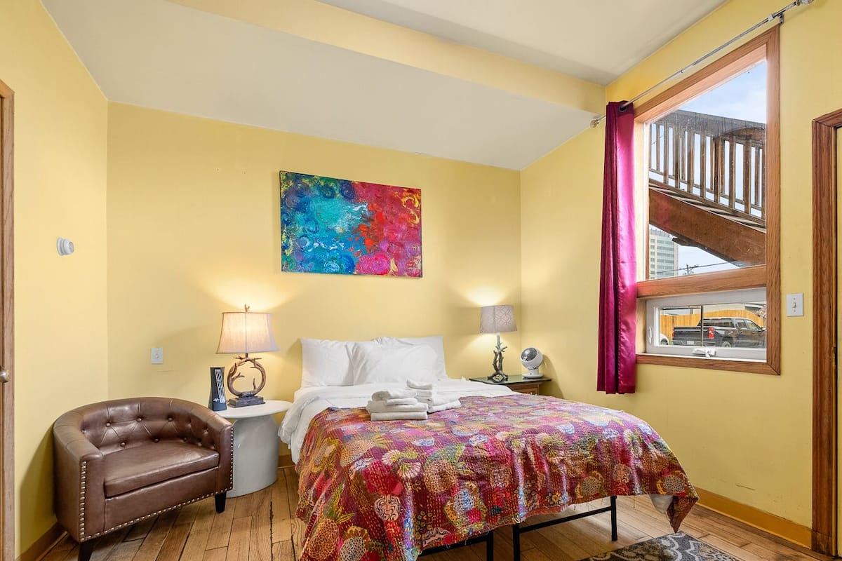 Un dormitorio con cama, dos mesitas de noche, una silla, una obra de arte colorida en la pared y una ventana con cortinas.