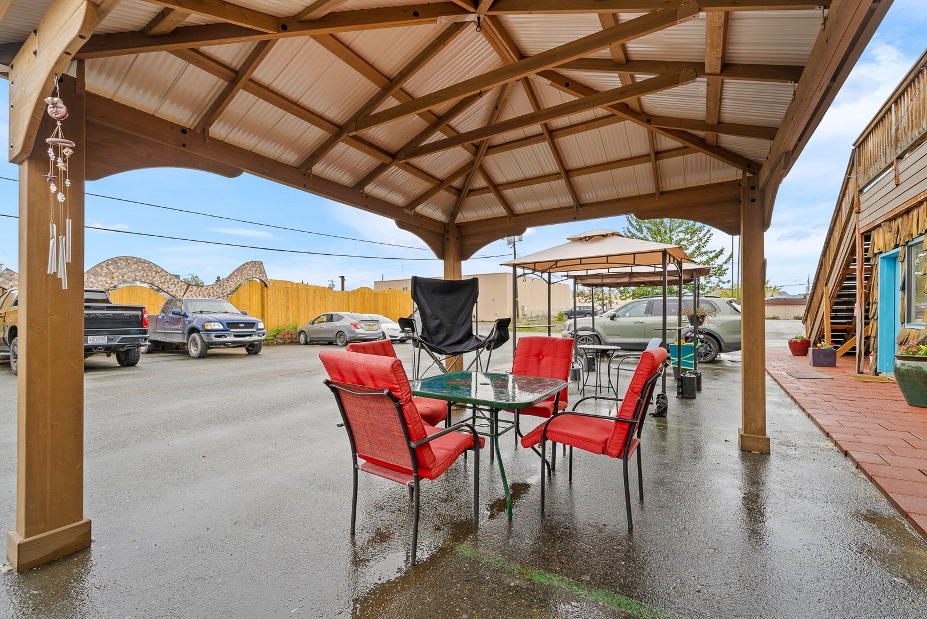 A covered outdoor seating area with a table and chairs, a wooden pergola, and a view of a street with parked cars.