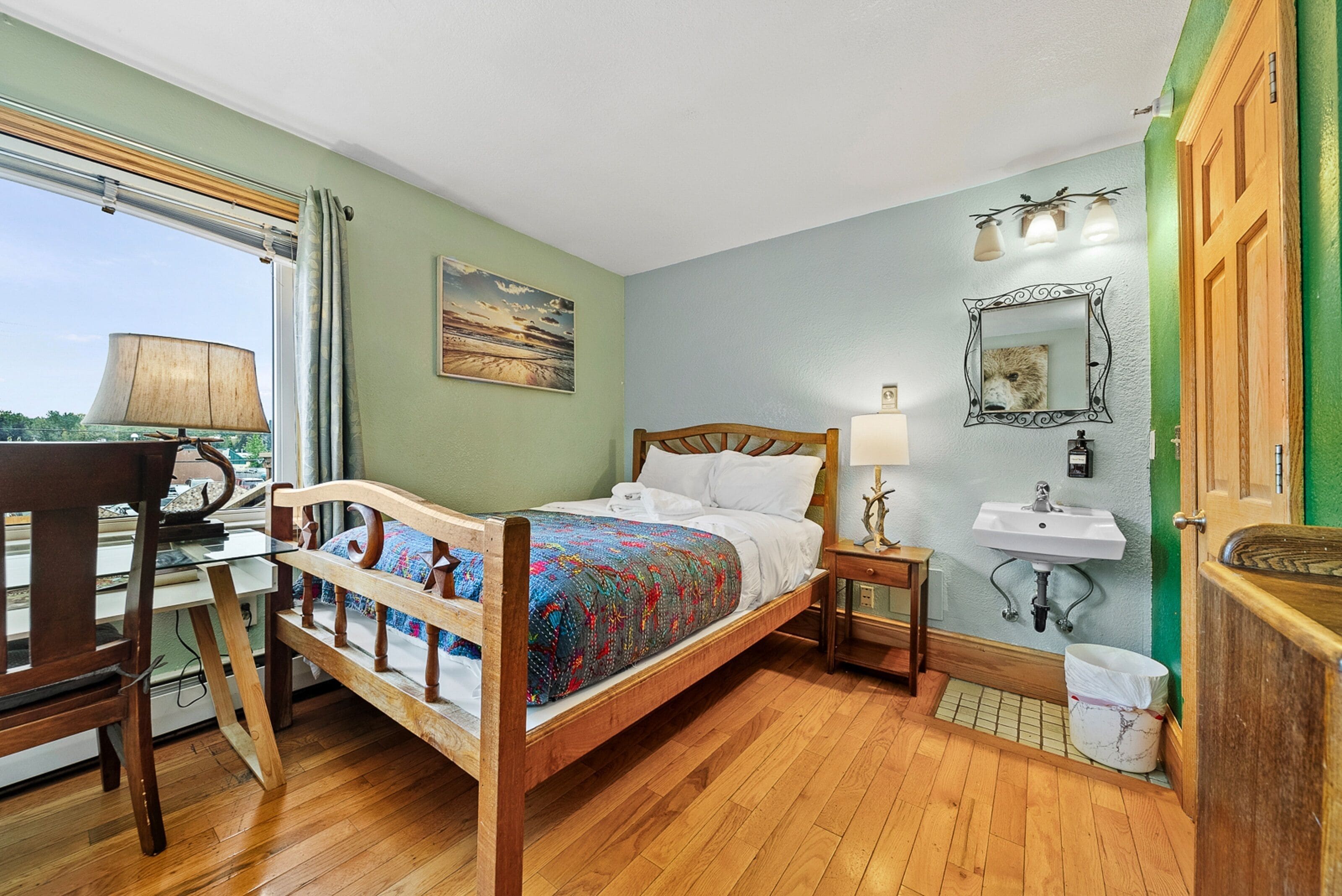 A bedroom with a wooden bed, a bedside table with a lamp, a window with curtains, a painting on the wall, and a green door.