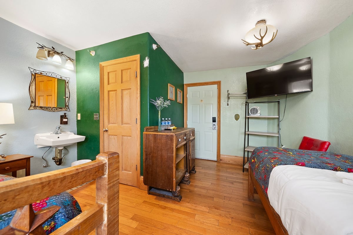 Una habitación con una cama de madera, un televisor montado en la pared, un aparador de madera, un espejo y un lavabo.