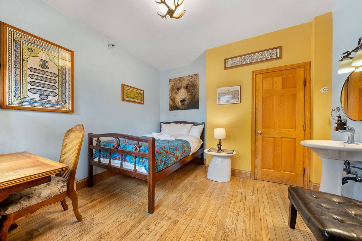 Una habitación con piso de madera, una cama con cubrecama colorido, una silla de madera, una mesita, una lámpara de pared y un espejo en la pared.