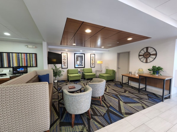 A modern hotel lobby with a sofa, green armchairs, a coffee table, a clock, and a TV.