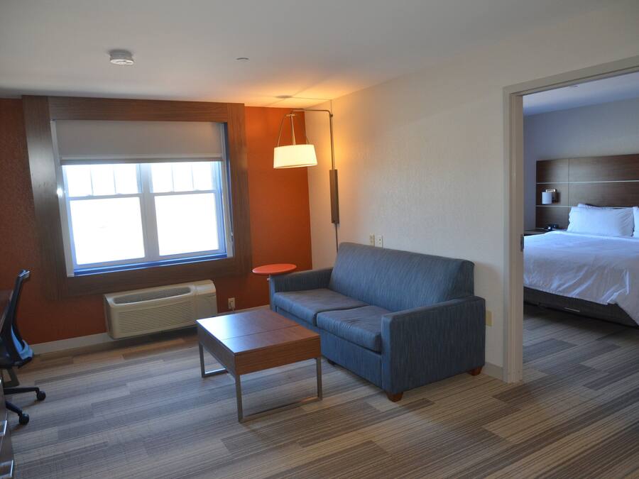Habitación de hotel con un ventanal grande, un sofá, una mesita, una lámpara y una cama con cabecera.
