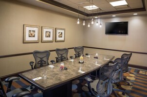 Un salón de conferencias con una mesa larga, seis sillas, un televisor en la pared y cuadros con motivos florales.