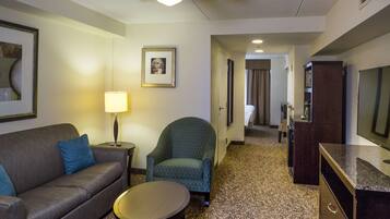 A hotel room with a sofa, armchair, small table, lamp, and a television.