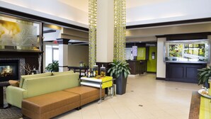 A modern lobby with a green sofa, a fireplace, a dining area, and a view of a garden through a window. - Hilton Garden Inn Hampton Coliseum Central (Hampton)