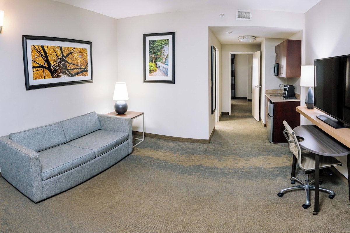 A hotel room with a grey sofa, a desk with a computer, a flat-screen TV, and framed pictures on the walls.