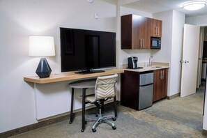 Habitación de hotel pequeña con kitchenette, escritorio, televisor de pantalla plana, una silla y una lámpara.