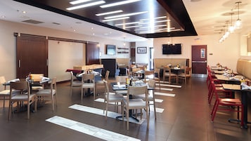 Un restaurante moderno con varias mesas y sillas, un televisor de pantalla plana en la pared y un techo con luminarias lineales.