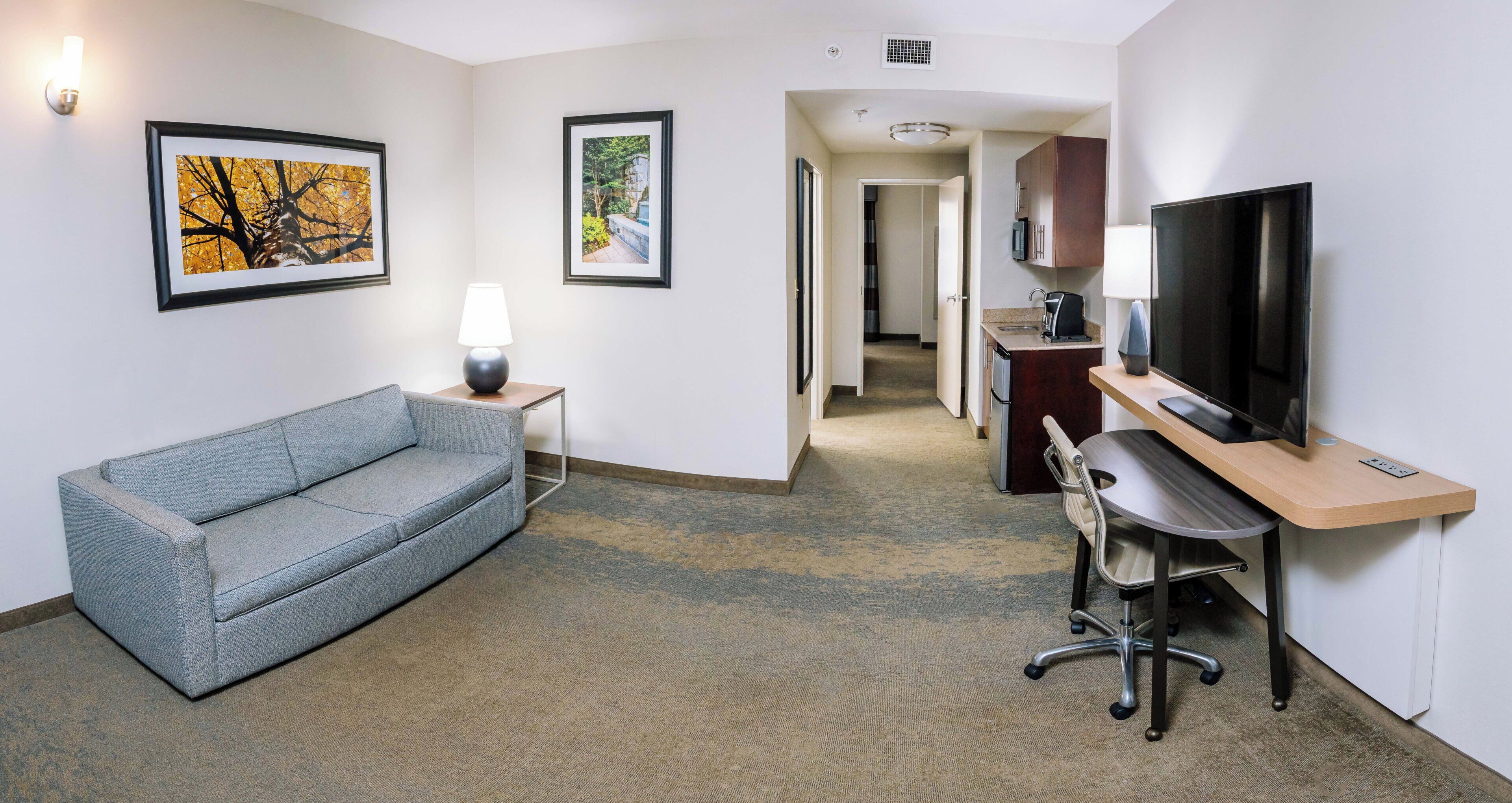 A hotel room with a grey sofa, a desk with a chair, a television, and framed pictures on the walls.
