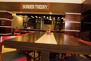 Interior de restaurante con una larga mesa de madera, sillas rojas y un letrero en la pared que dice 'BURGER THEORY'.