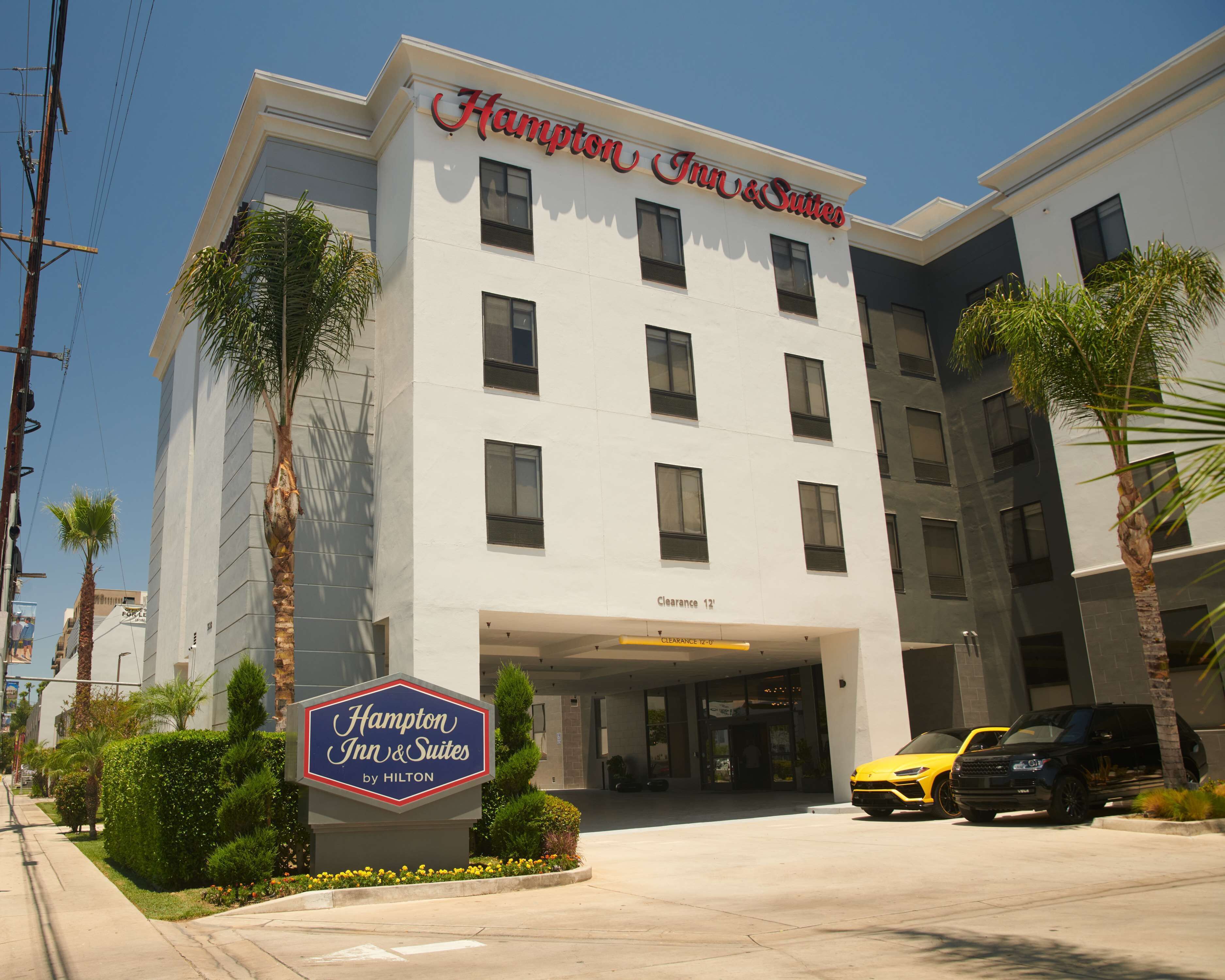 Edificio di un Hampton Inn & Suites by Hilton con ingresso ben visibile, palme e un'auto gialla parcheggiata davanti.