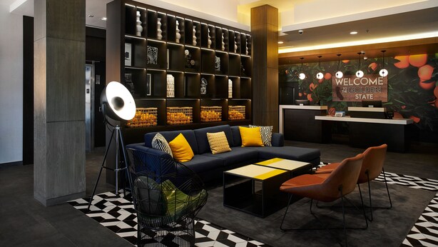 A modern lobby with a sofa, chairs, a coffee table, a bookshelf, and a reception desk. - Hampton Inn & Suites Los Angeles/Sherman Oaks (Van Nuys)