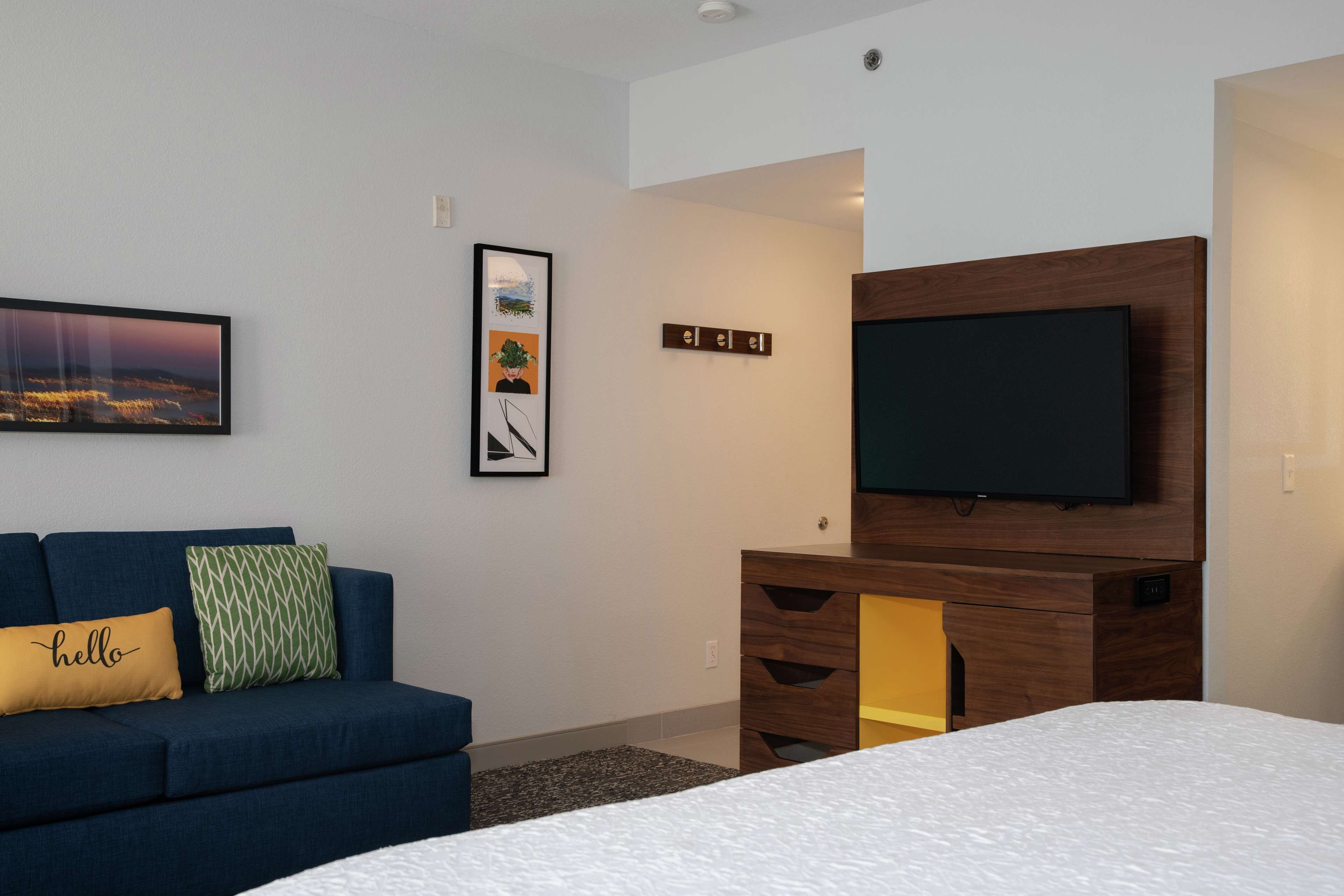 A modern hotel room with a large bed, a blue sofa with decorative pillows, a flat-screen TV mounted on a wooden cabinet, and a framed picture on the wall.