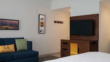 A modern hotel room with a large bed, a blue sofa with decorative pillows, a flat-screen TV mounted on a wooden cabinet, and a framed picture on the wall.