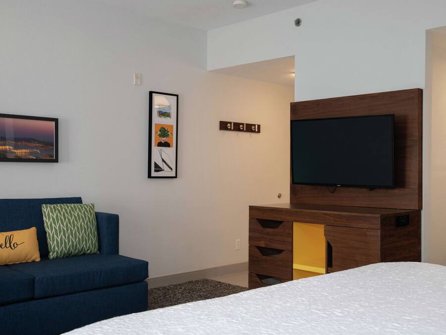 A modern hotel room with a large bed, a blue sofa with decorative pillows, a flat-screen TV mounted on a wooden cabinet, and a framed picture on the wall.