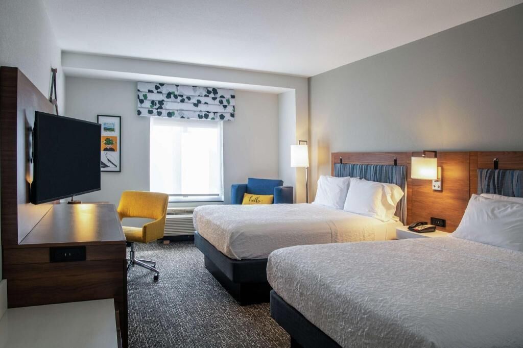 A hotel room with two beds, a desk with a TV, a chair, and a window with curtains.