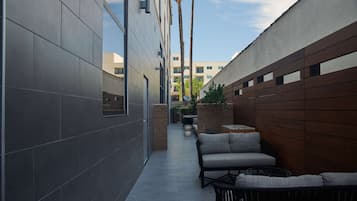 A narrow outdoor corridor with a modern building on one side and a wooden wall on the other. There are outdoor seating areas with cushions and tables.