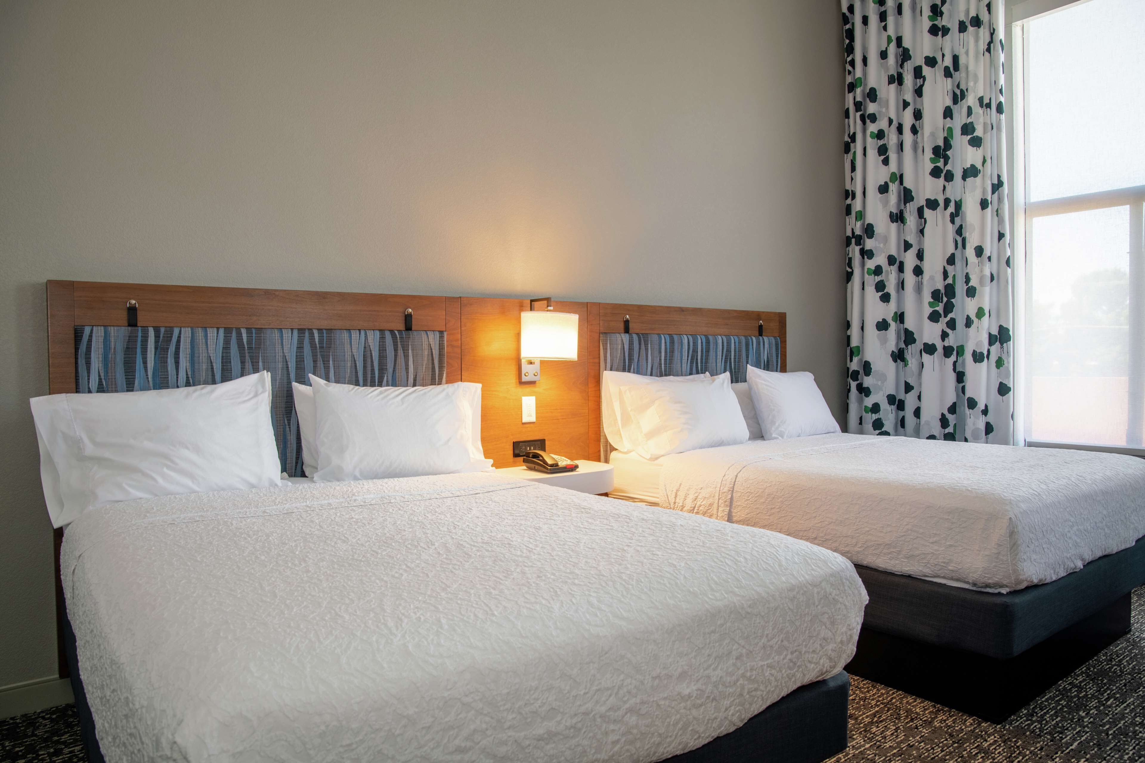 Una habitación de hotel con dos camas, una mesita de noche con lámpara y una ventana con cortinas.