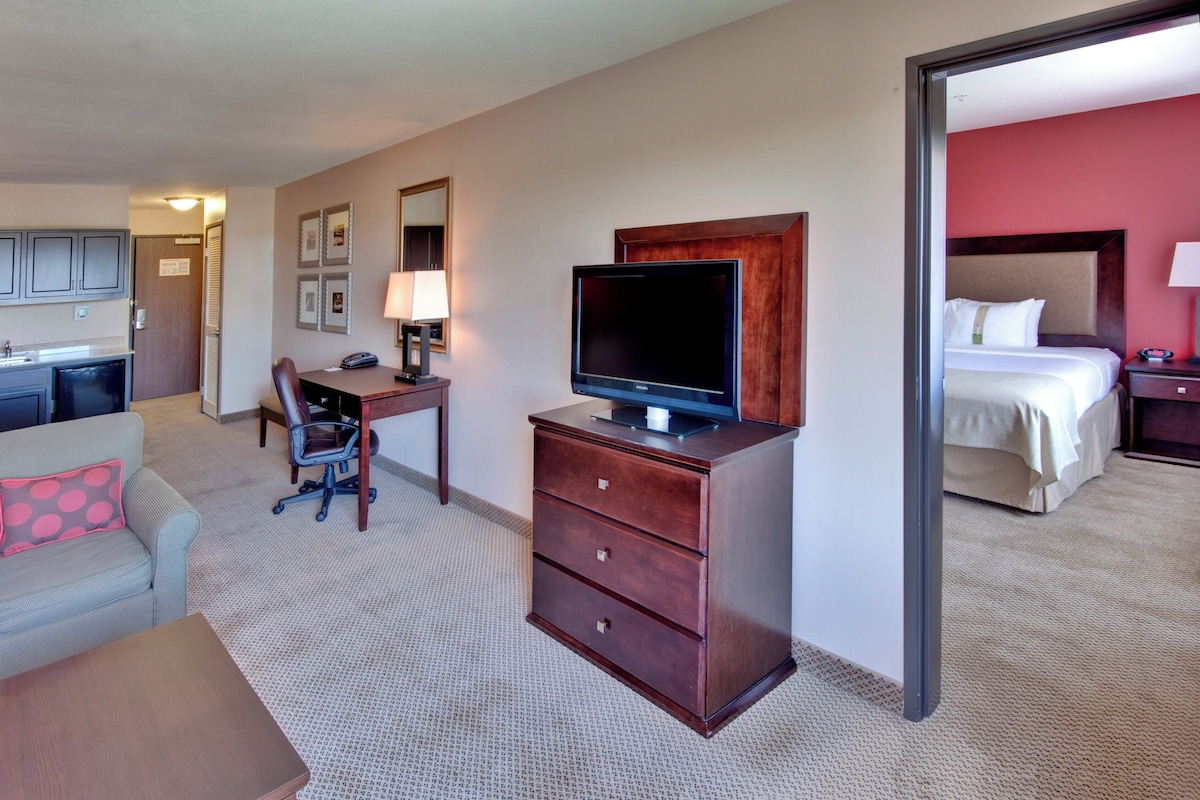 Una habitación de hotel con kitchenette, un televisor sobre un mueble de madera, un escritorio con una silla y una cama con cabecera.