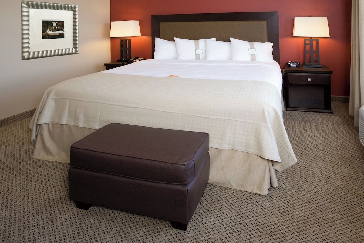Una habitación de hotel con una cama grande, dos mesitas de noche con lámparas, un cuadro enmarcado en la pared y un reposapiés marrón.
