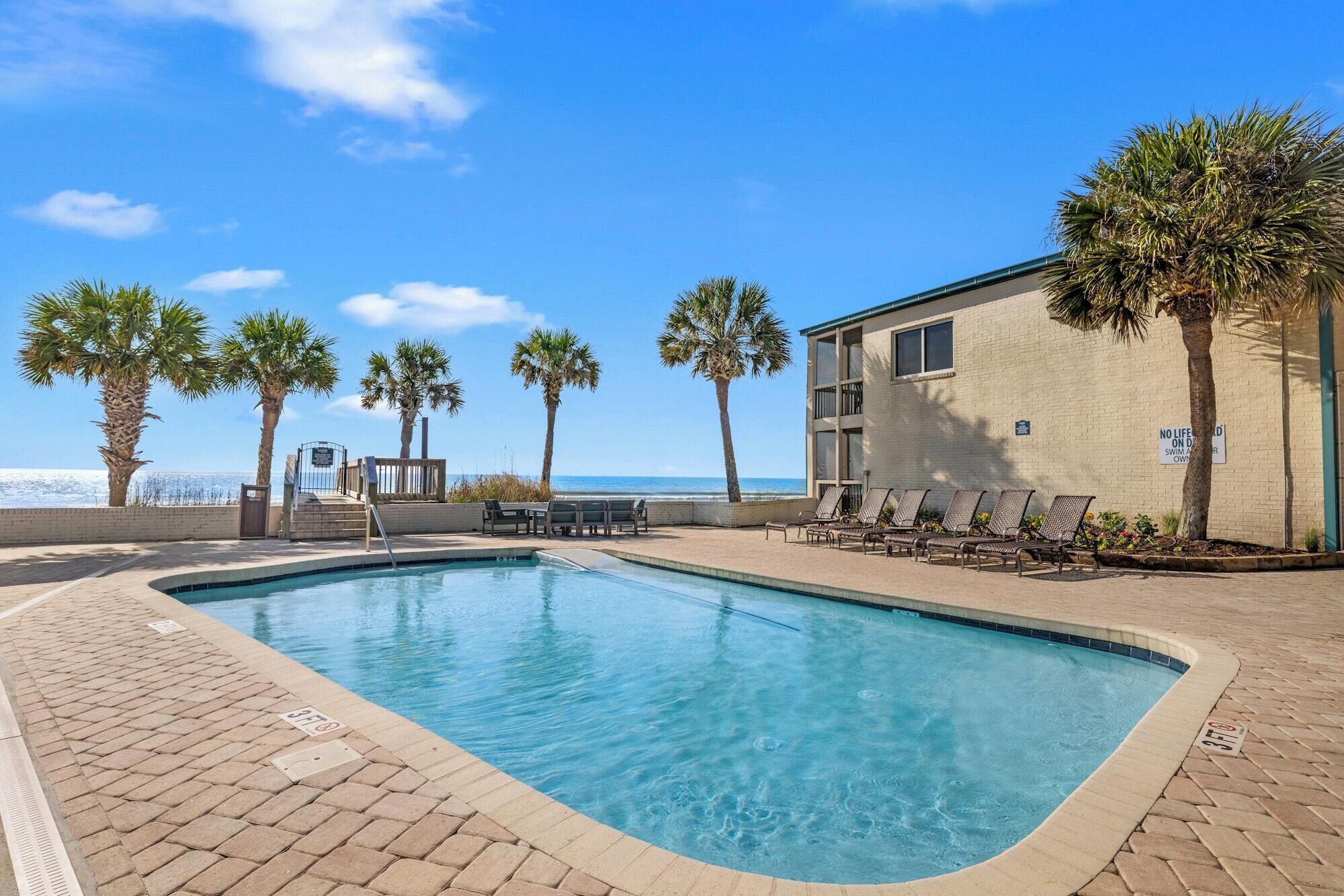A swimming pool with lounge chairs and palm trees, a building with a sign, and a clear sky.