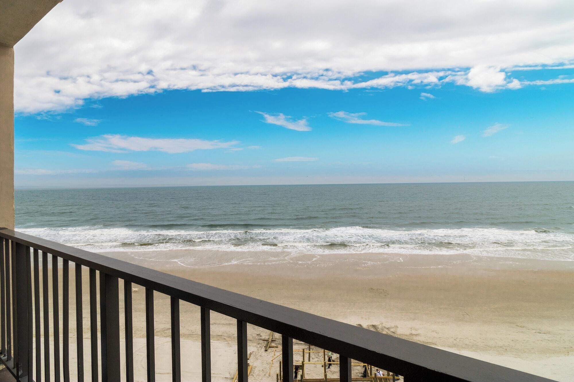 Vue sur la plage depuis un balcon doté d’une balustrade, avec l’océan en toile de fond et un ciel partiellement nuageux.