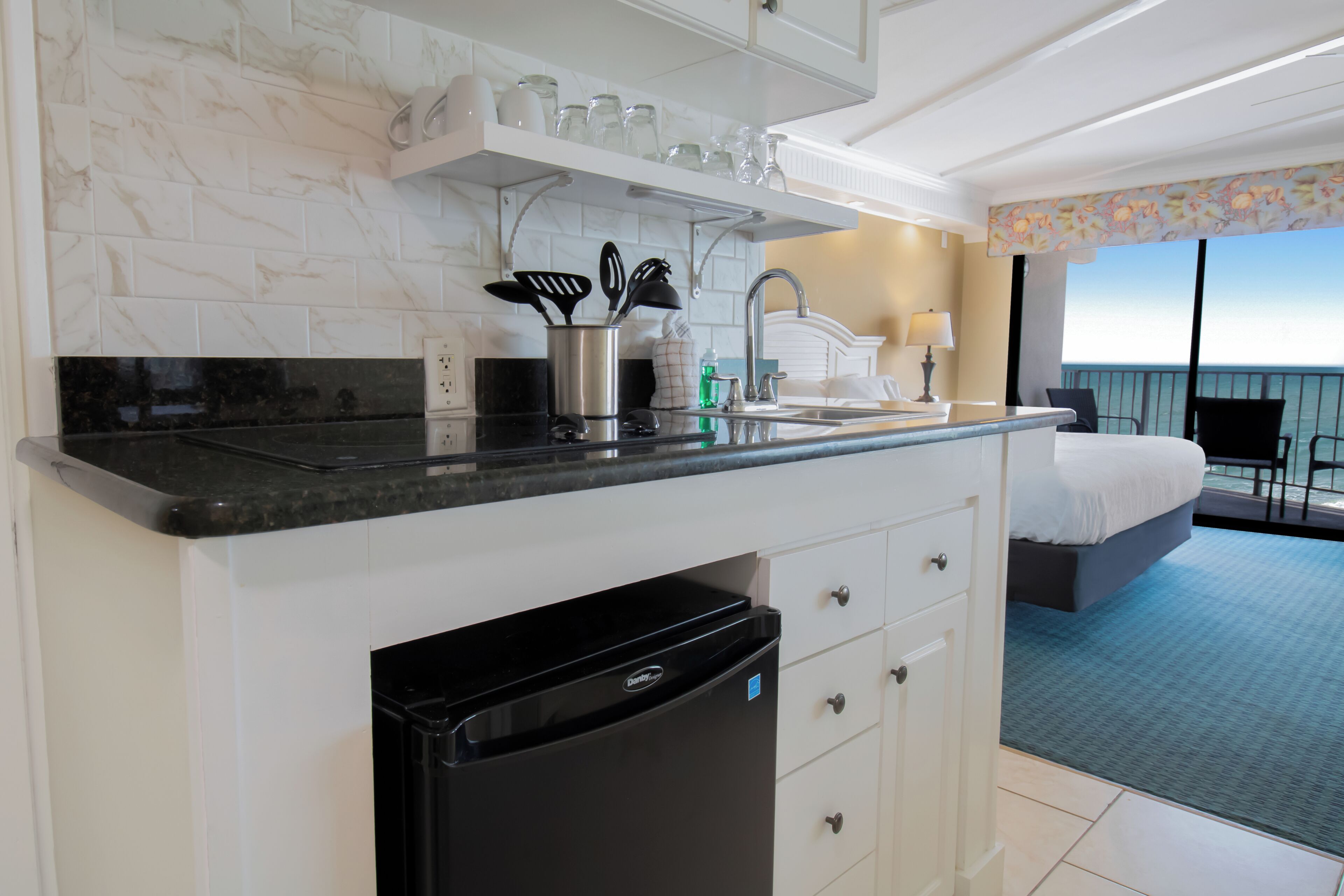 A kitchen with a dishwasher, a countertop, and a sink. There is a bed with a view of the ocean in the background.