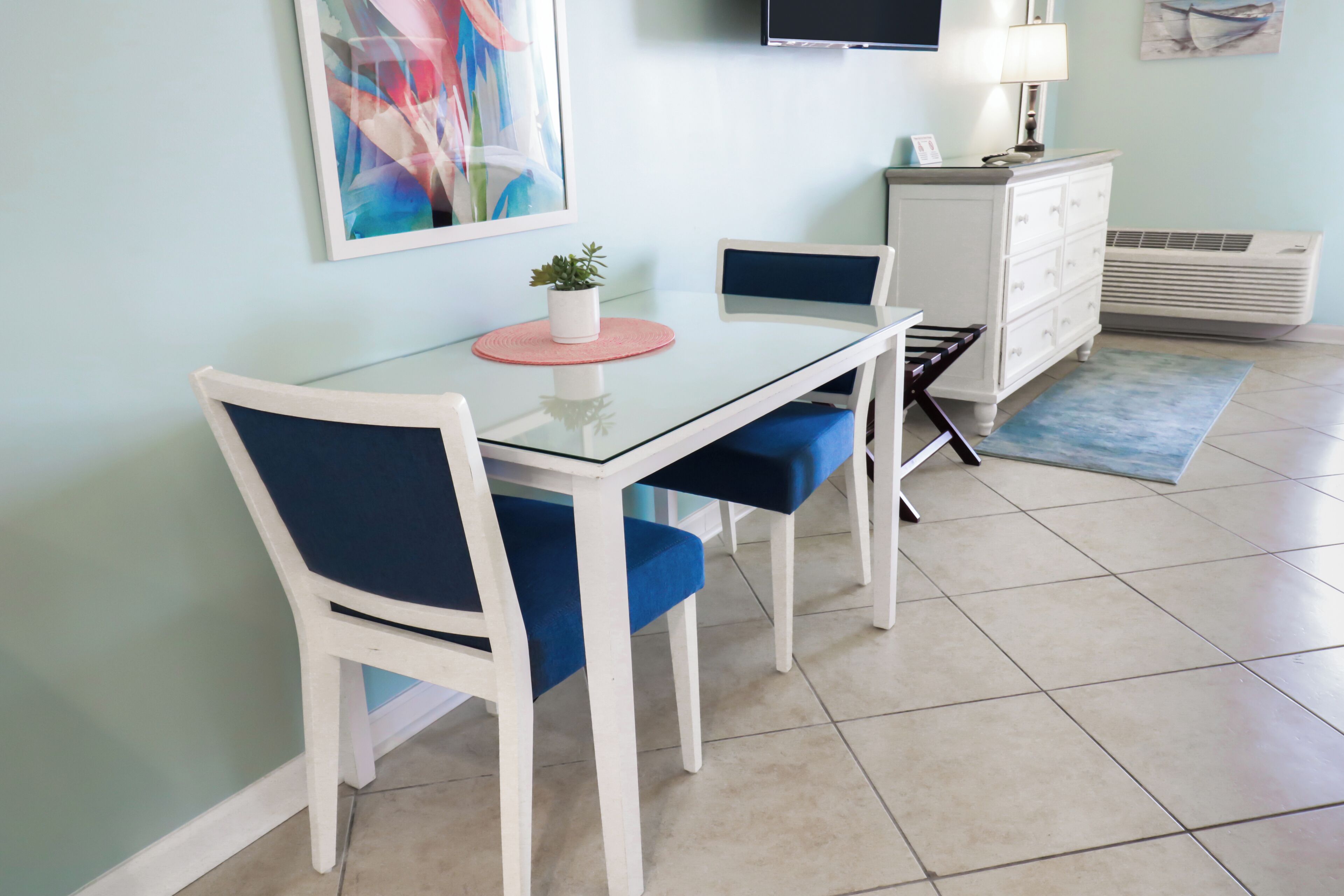 A dining area with a glass table, white chairs, a small potted plant, and a blue rug.