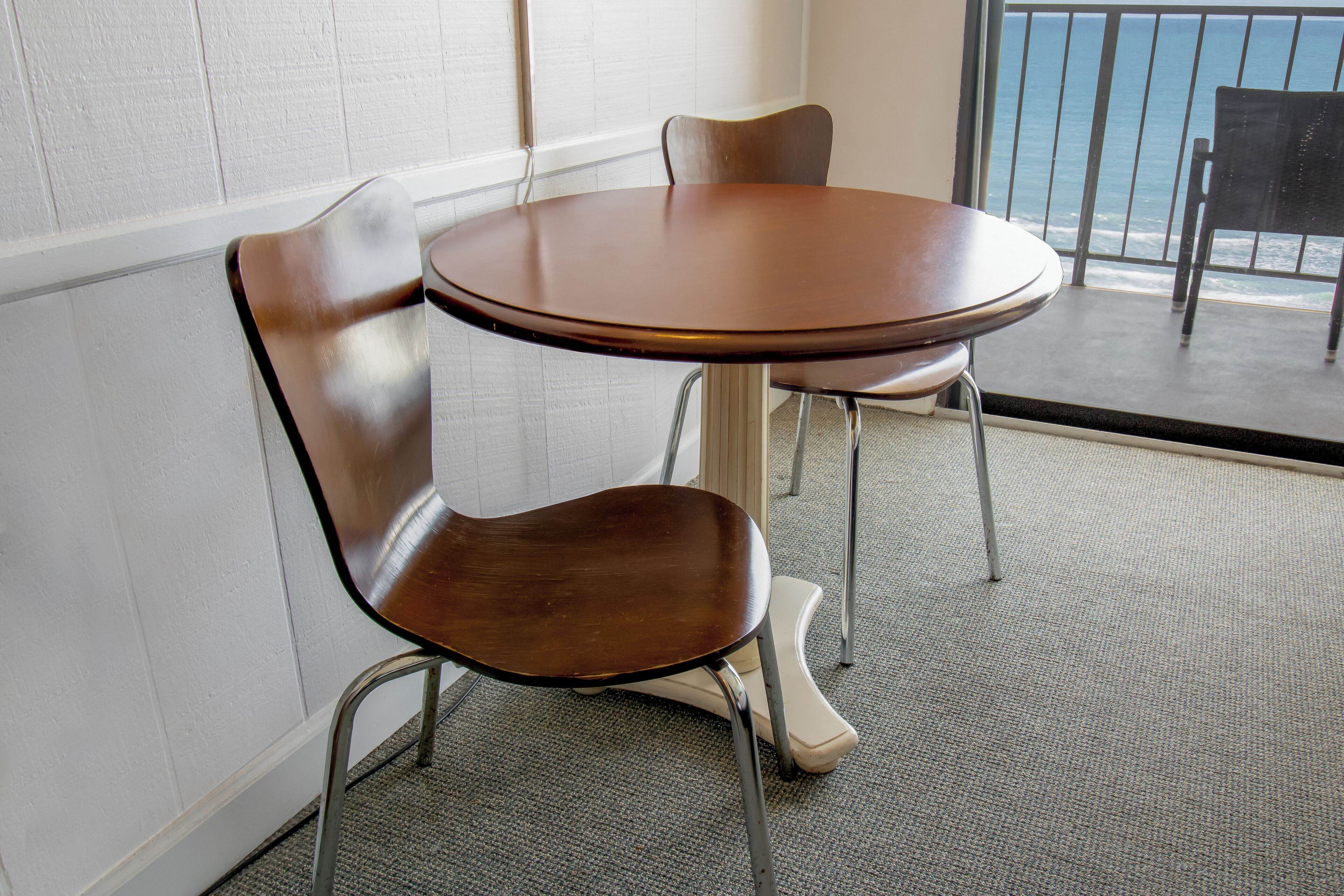 A round table with two chairs, a view of the ocean through a window, and a white wall.