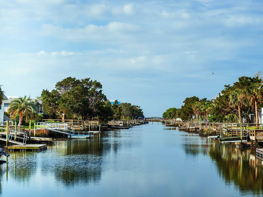Un río tranquilo con barcos amarrados a sus orillas, rodeado de árboles y un puente a lo lejos.