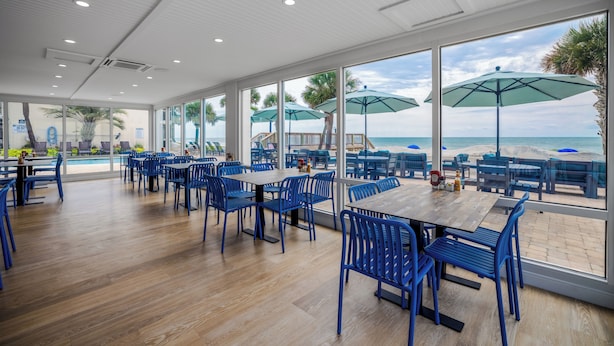 A spacious indoor dining area with large windows overlooking a beach, wooden flooring, and blue chairs and tables. - The Oceanfront Litchfield Inn (Pawleys Island)