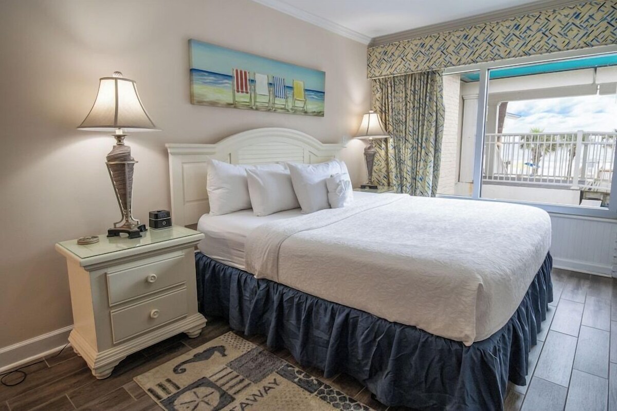 A bedroom with a bed, two bedside lamps, a nightstand, a painting, a patterned curtain, and a window with a view.