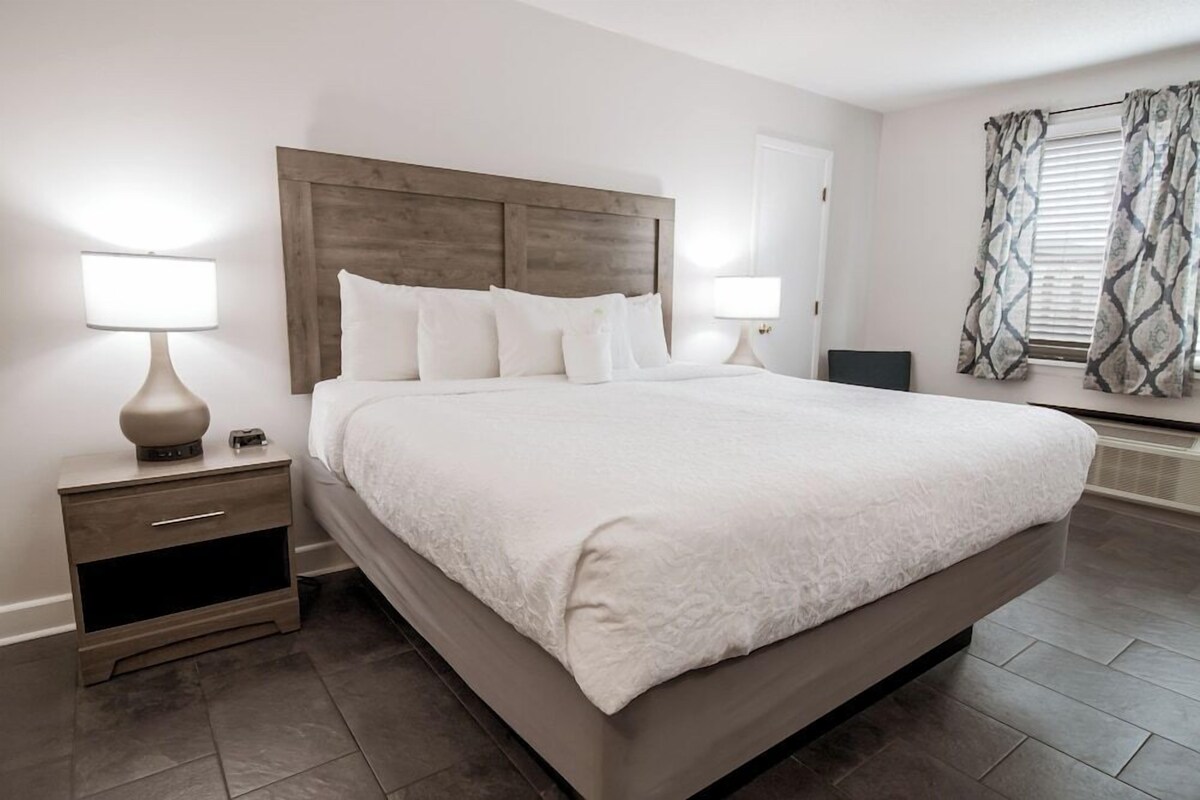 Un dormitorio moderno con una cama grande, dos mesitas de noche con lámparas, un cabecero de madera y una ventana con cortinas estampadas.