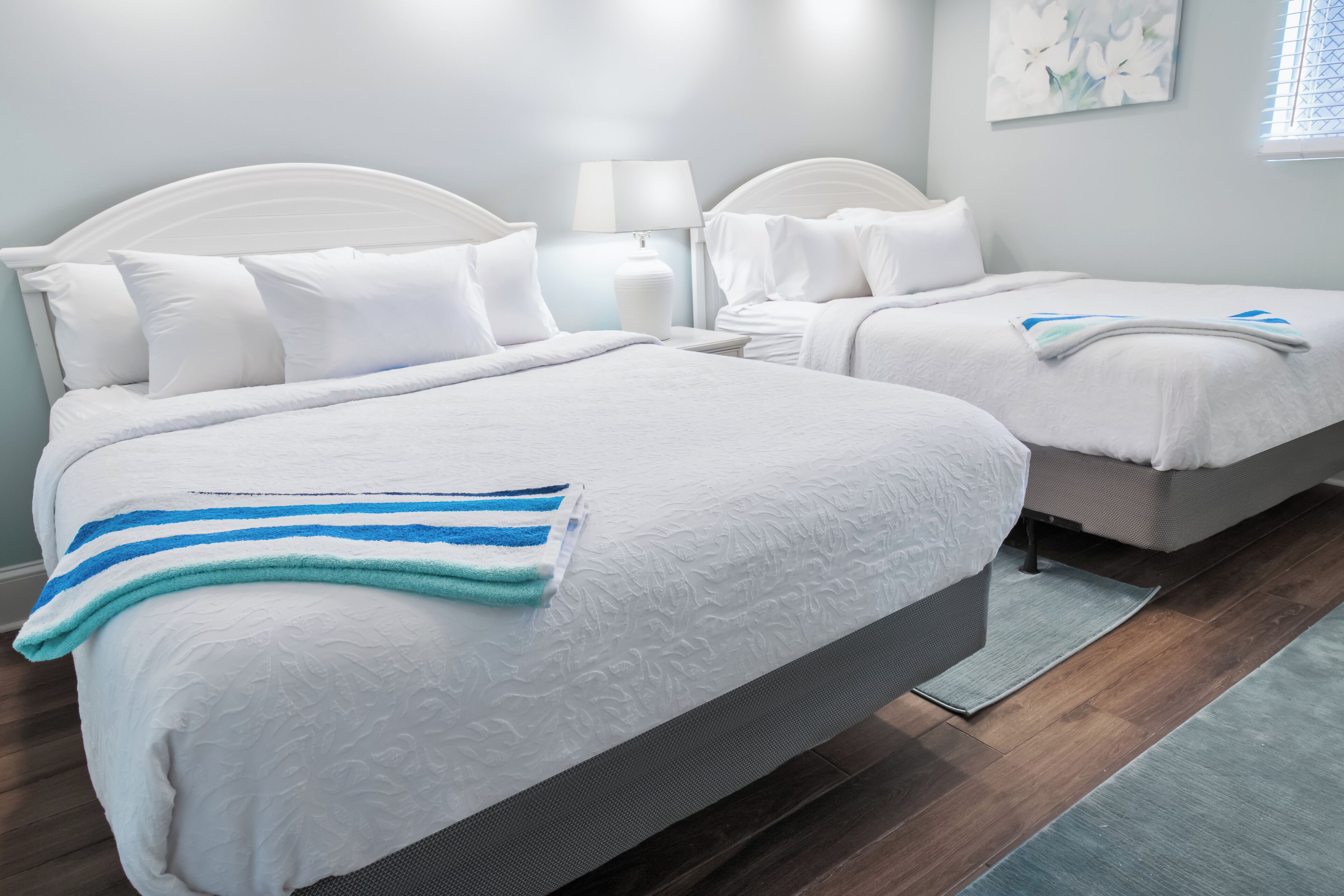 Una habitación de hotel con dos camas, ropa de cama blanca, una toalla a rayas, una mesita de noche con una lámpara y un cuadro en la pared.