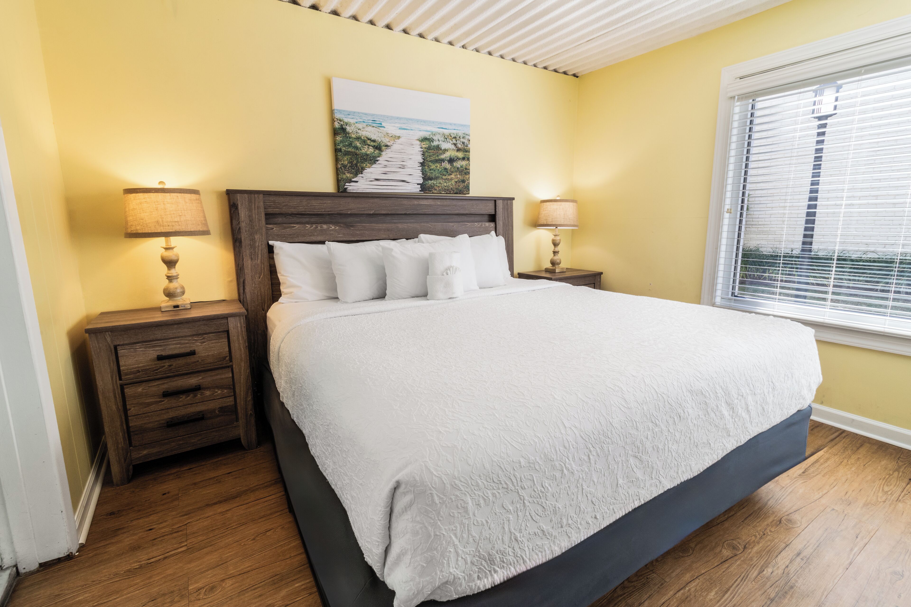 A bedroom with a large bed, wooden headboard, two bedside tables with lamps, a painting on the wall, and a window with blinds.