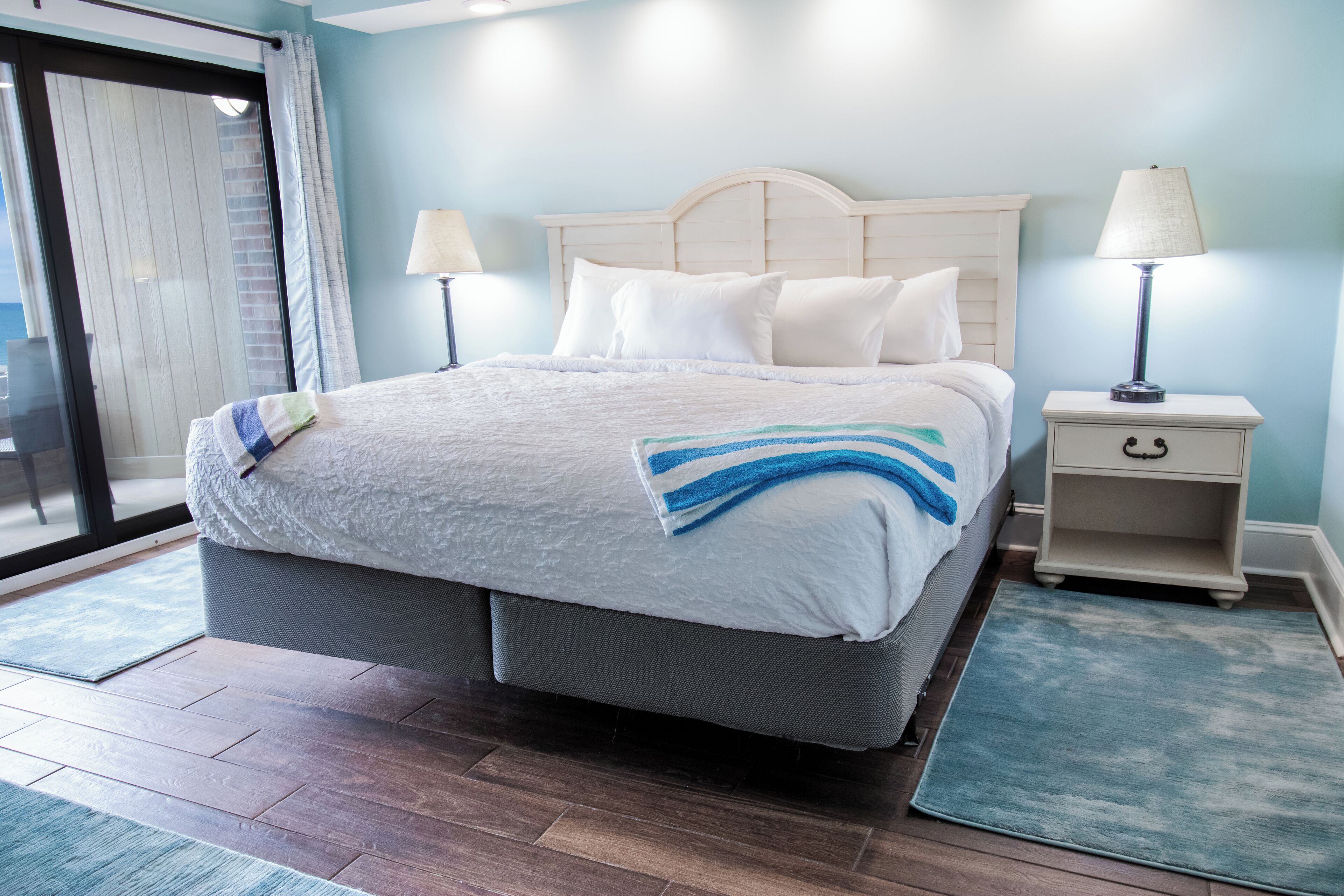 A neatly made bed with a white and blue striped blanket, a bedside table with a lamp, and a view of a balcony through a glass door.