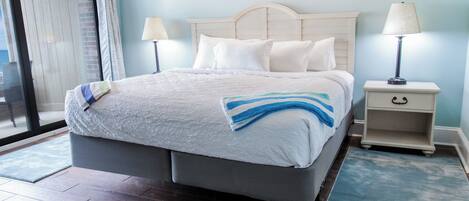 A neatly made bed with a white and blue striped blanket, a bedside table with a lamp, and a view of a balcony through a glass door.
