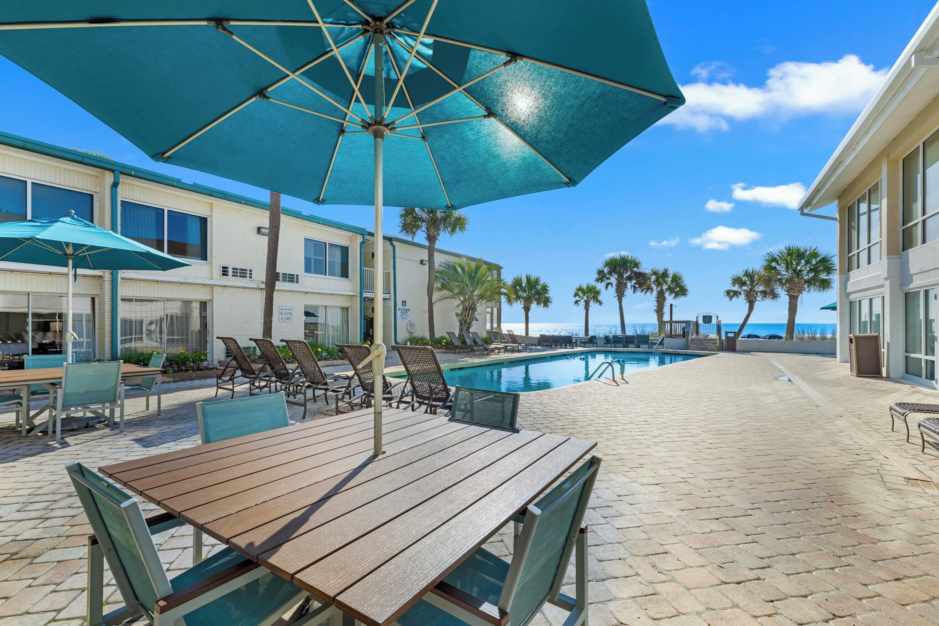 A poolside area with a wooden table, chairs, and a large umbrella.