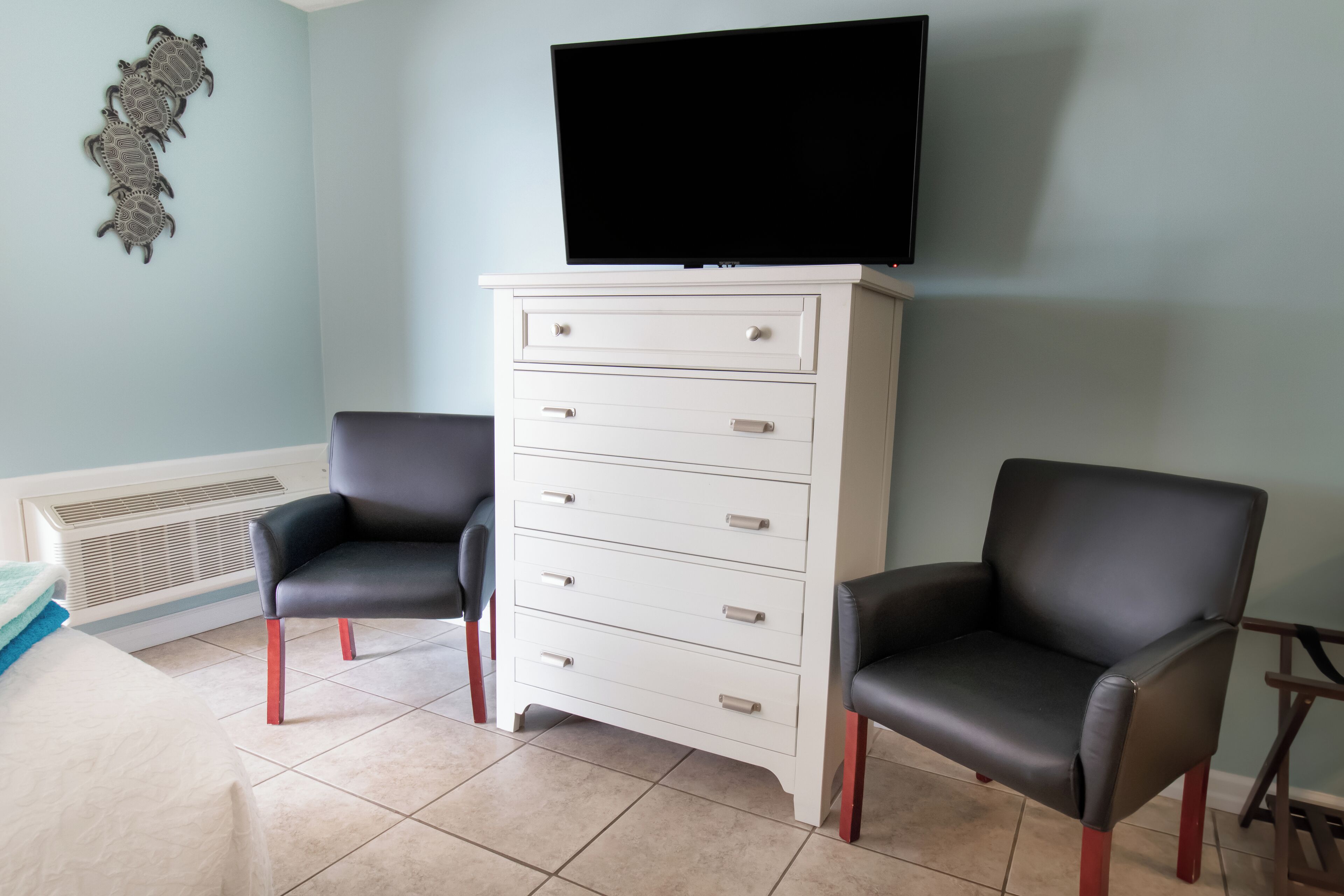 A room with two black leather chairs, a white dresser with drawers, a flat-screen TV mounted above the dresser, and a decorative wall piece.