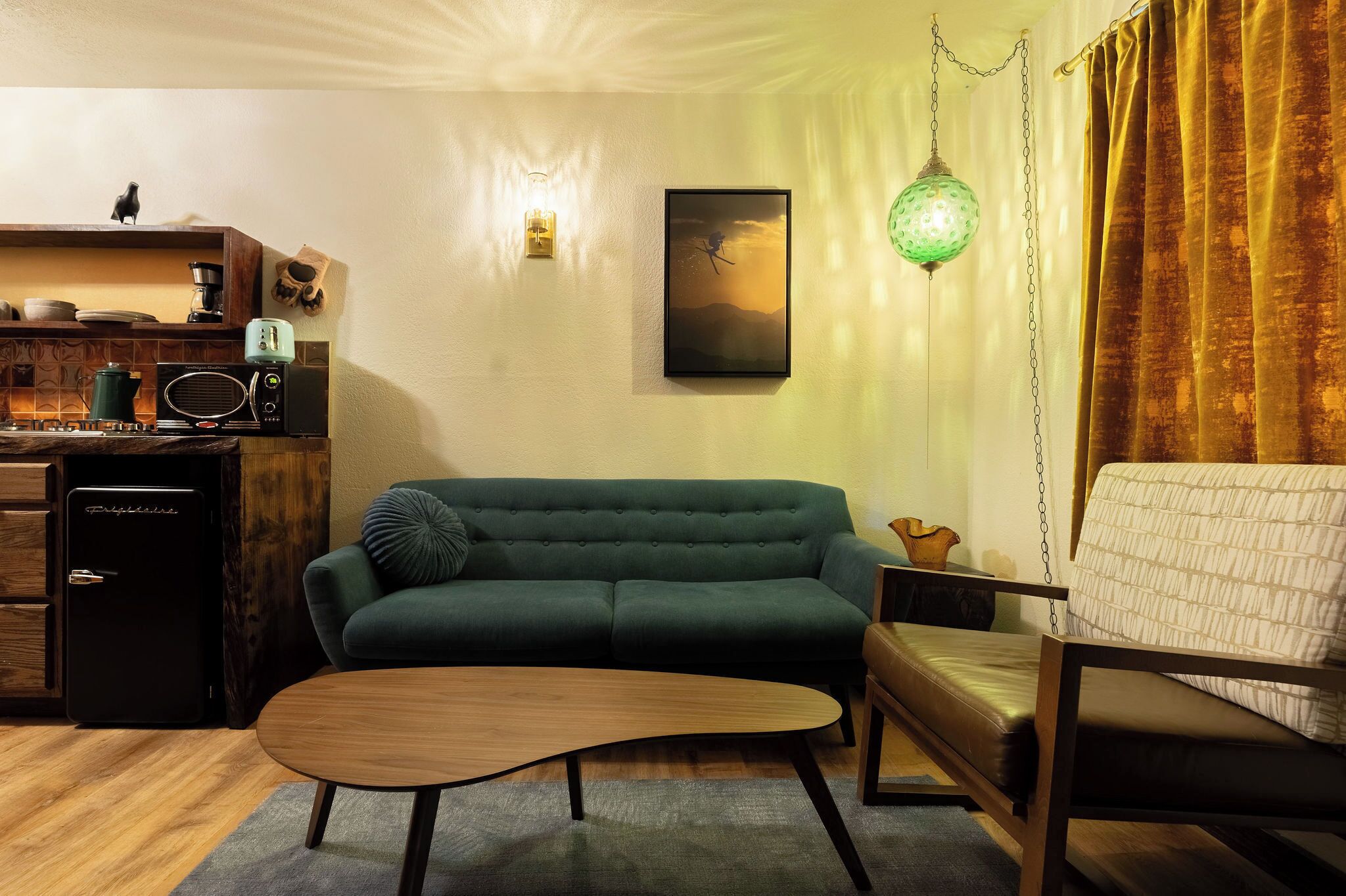 Een gezellige woonkamer met een groene bank, een houten salontafel en een kleine keukenhoek met een koelkast en een radio.