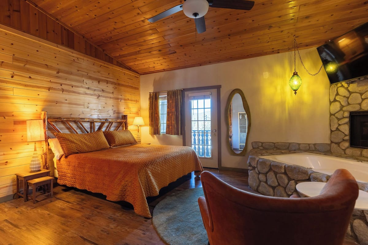 A cozy wooden cabin bedroom with a bed, a stone fireplace, a bathtub, and a ceiling fan.