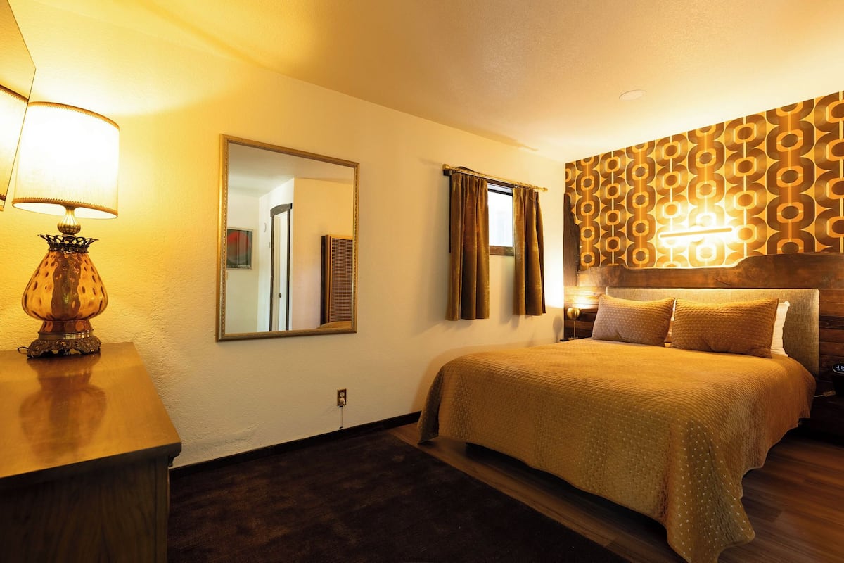 A hotel room with a large bed, a bedside table with a lamp, a mirror, and a patterned wall.