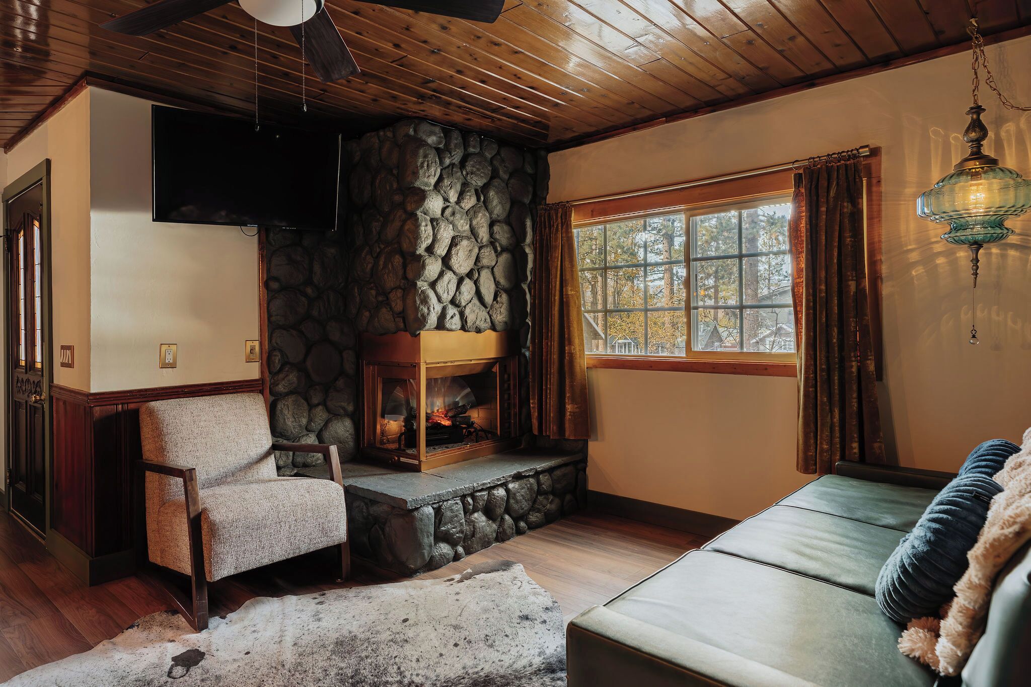 A cozy living room with a stone fireplace, a comfortable sofa, a plush armchair, and a large window with curtains.