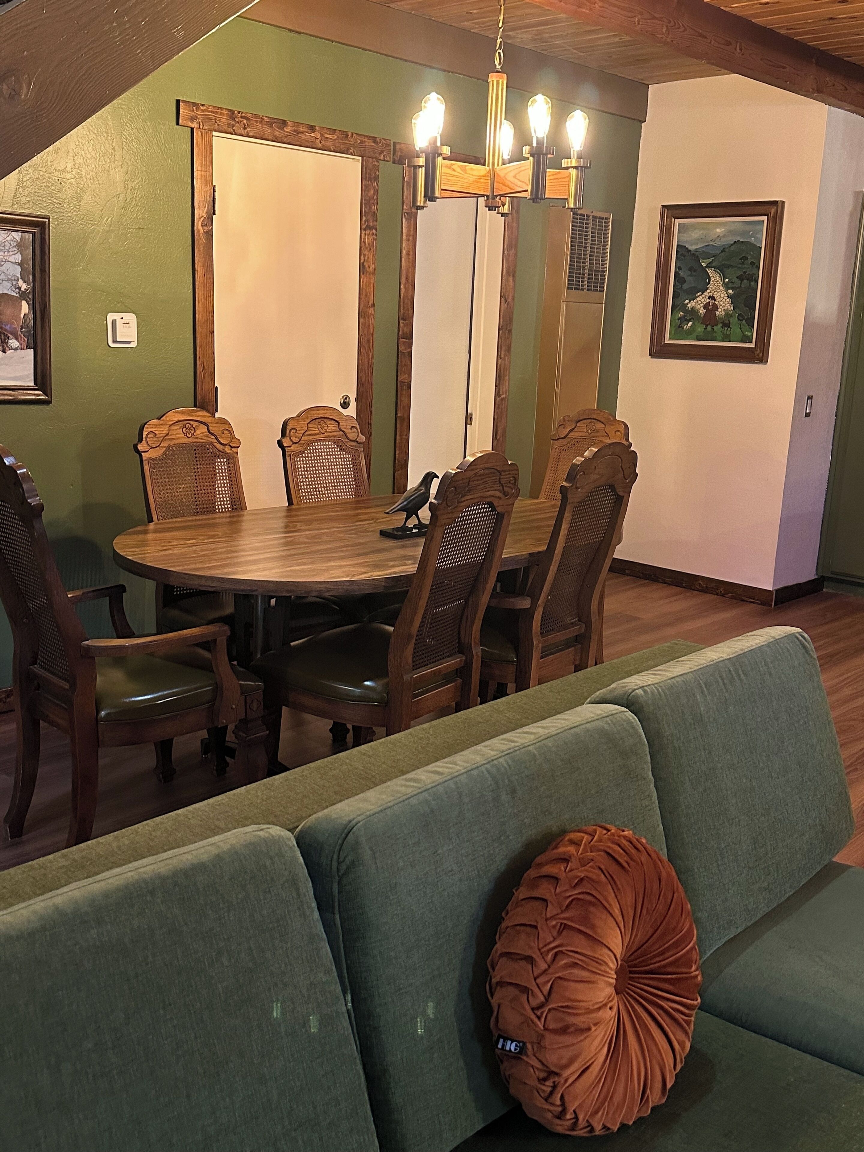 A dining area with a wooden table and chairs, a green sofa with a brown cushion, and a chandelier.